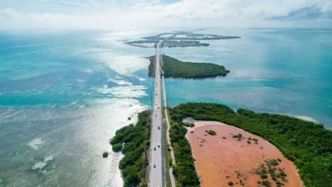 Scenic aerial view of the Overseas Highway stretching across the turquoise waters of the Florida Keys, with lush green islands and a red salt pond in the foreground.
