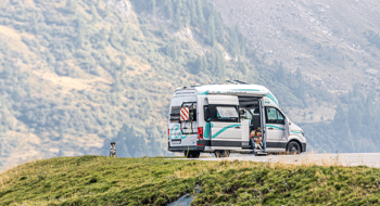 big size camper from roadsurfer standing on a parking in the mountains with a family inside and the dog waiting outside