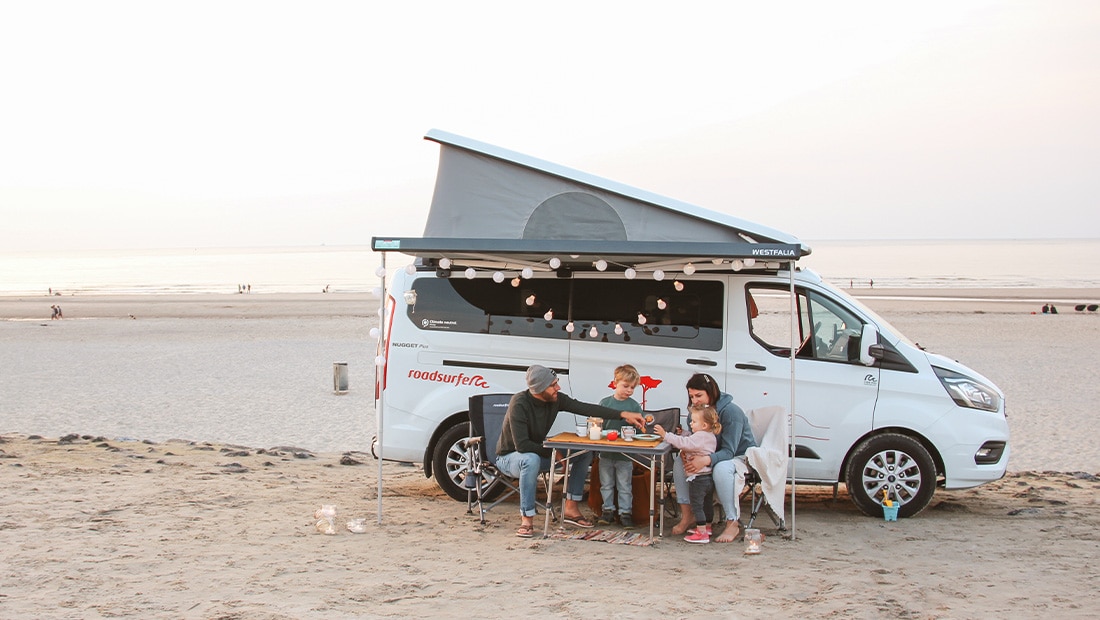 Family of four sitting at a table in front of a Ford Nugget parked on the beach