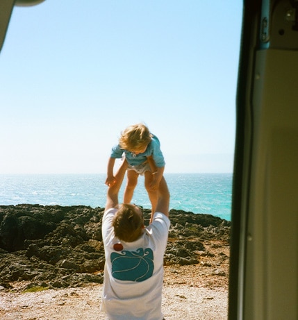 Joyful moment of a man lifting a young child into the air near a rocky coastline, with the ocean and clear blue sky in the background