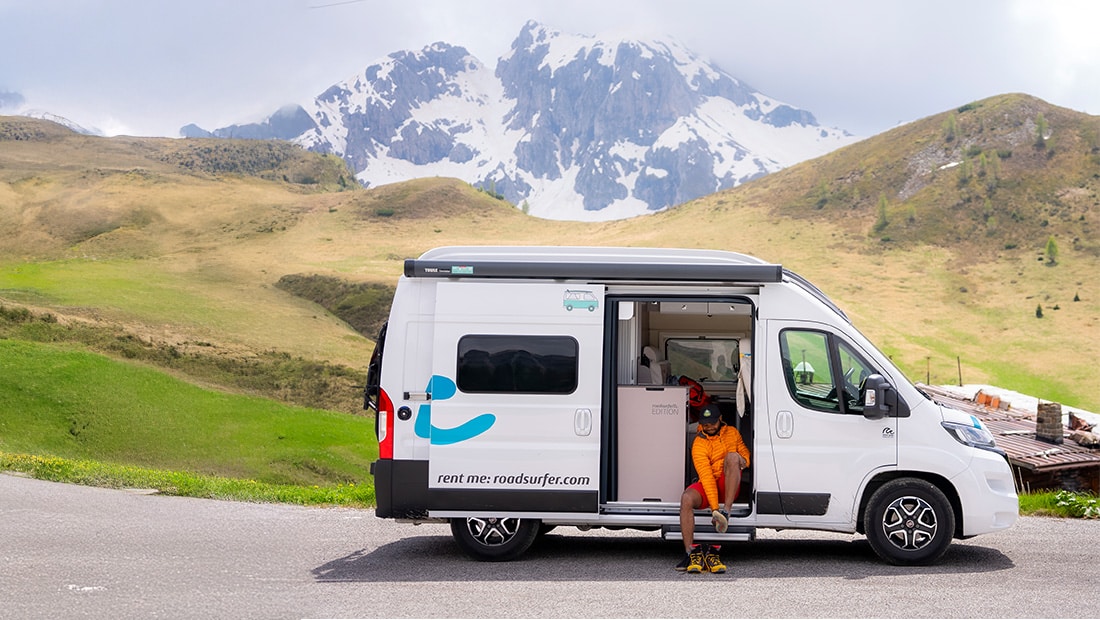 Man sitting in the open doorf of a campervan which is parked in the mountains