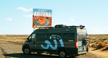 Grey roadsurfer boxvan photograph standing at the roadside in front of a Arizona state sign.
