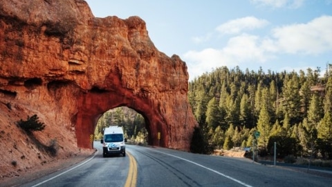 Roadsurfer camper van driving through a red rock tunnel surrounded by pine trees in Utah.