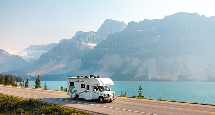 Scenic mountain landscape with a white roadsurfer RV driving along a highway beside a turquoise lake, surrounded by lush greenery and towering peaks with patches of snow