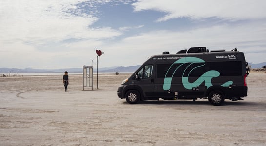 RV parked in the desert and a woman walks up to a phone booth with a love heart sculpture